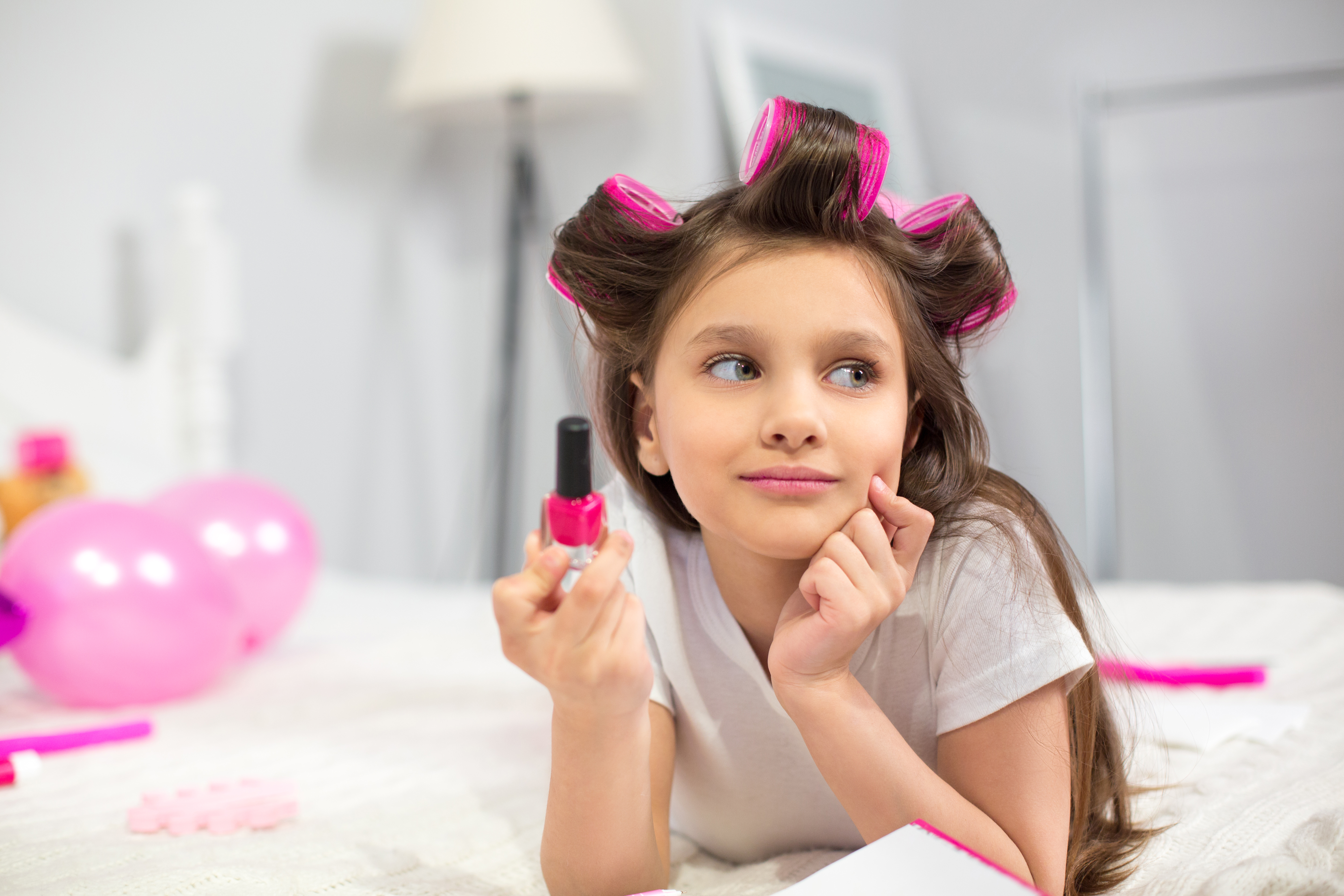Cute preschooler laying on white blanket holding nail polish. Lovely looking girlie with hair rollers looking at side holding bottle of pink nail polish in her hand.