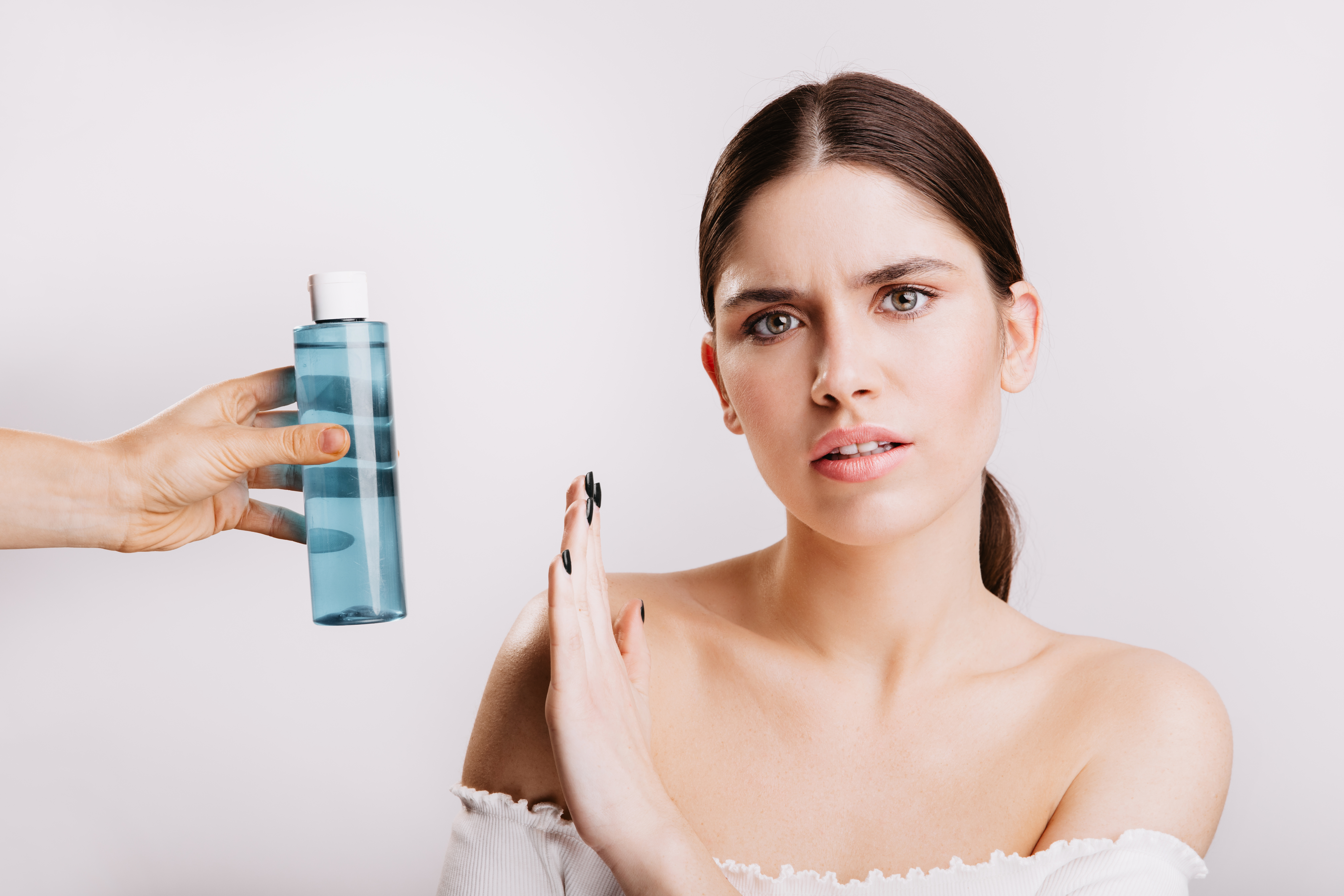 Portrait of girl with displeased facial expression on white background with micellar water. Woman without makeup against use of cosmetics